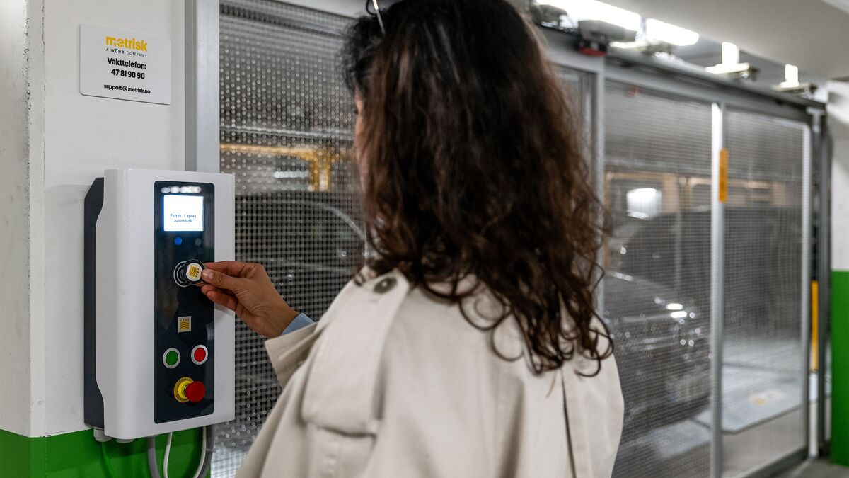 En person använder kontrollpanelen för ett halvautomatiskt parkeringssystem för att hämta ett parkerat fordon. I bakgrunden syns de flyttbara parkeringsplatserna och ett fordon inne i parkeringsanläggningen.